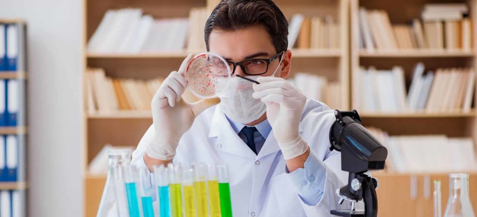 A person in laboratory clothing examining a dish beside a microscope