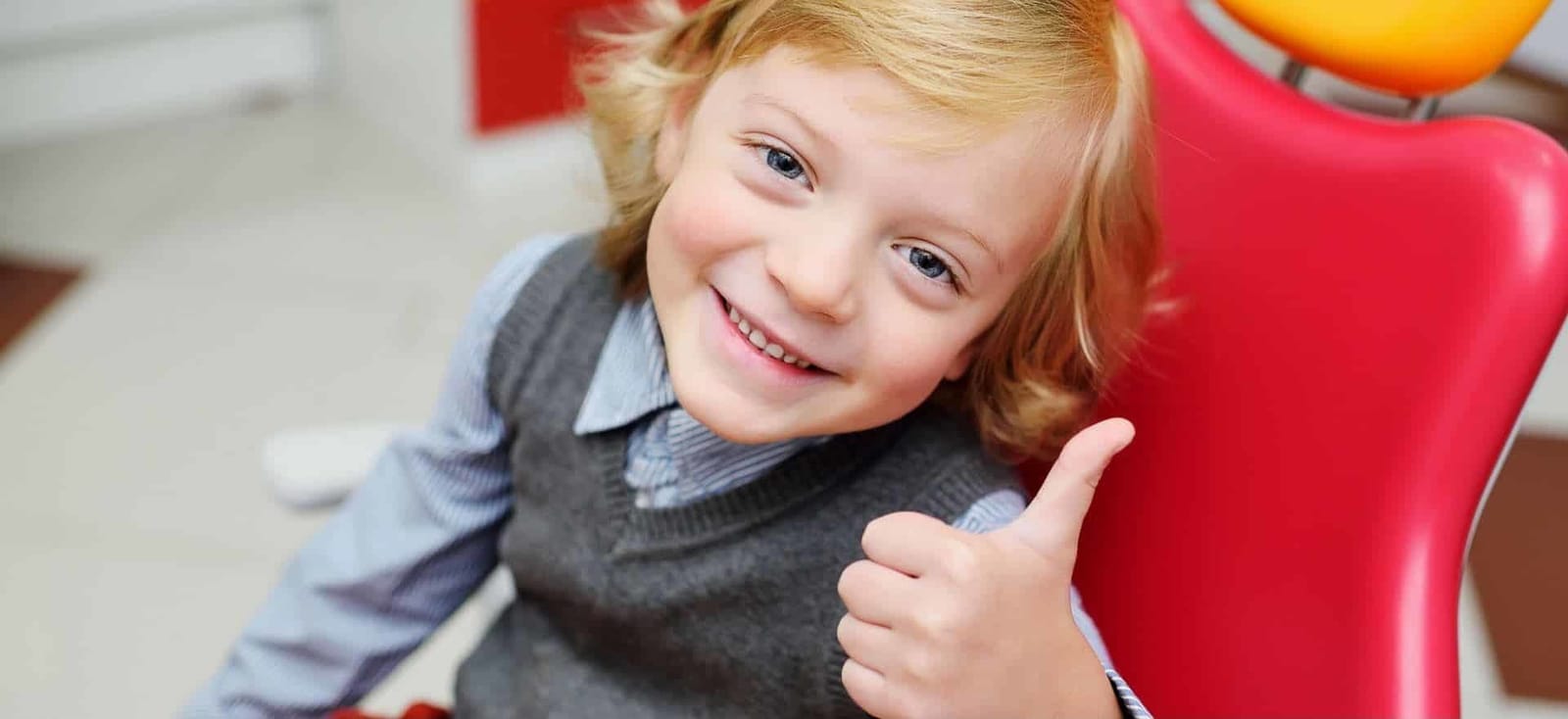 A smiling child giving a thumbs-up beside a red dental chair.