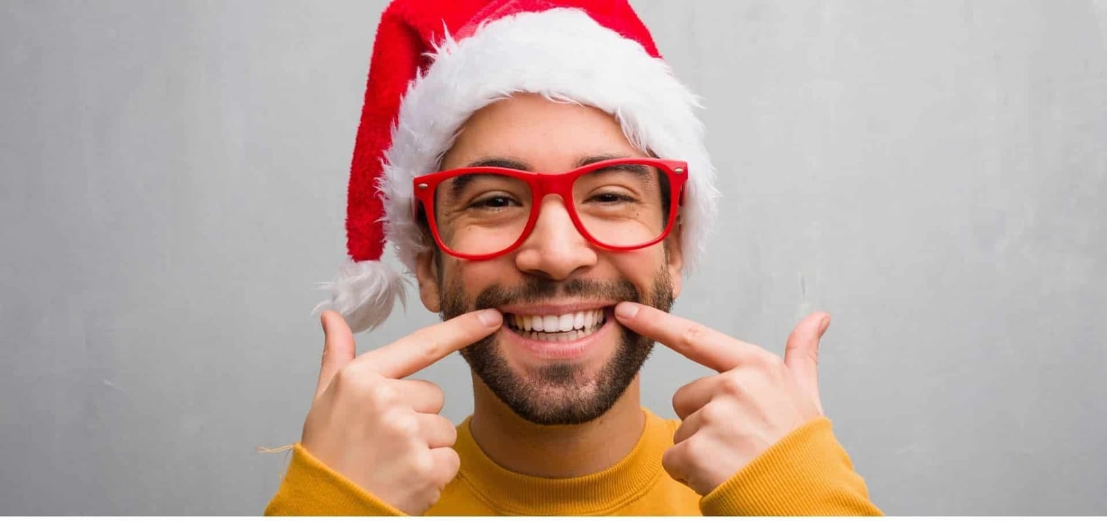 A smiling man in a Santa hat pointing towards his teeth