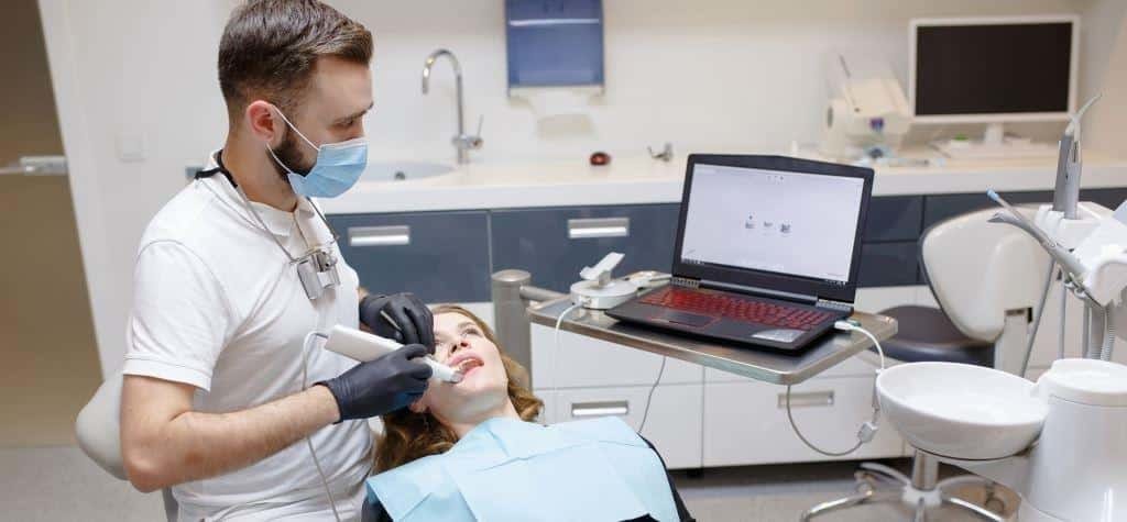 A dentist using a handheld scanner beside a patient and laptop