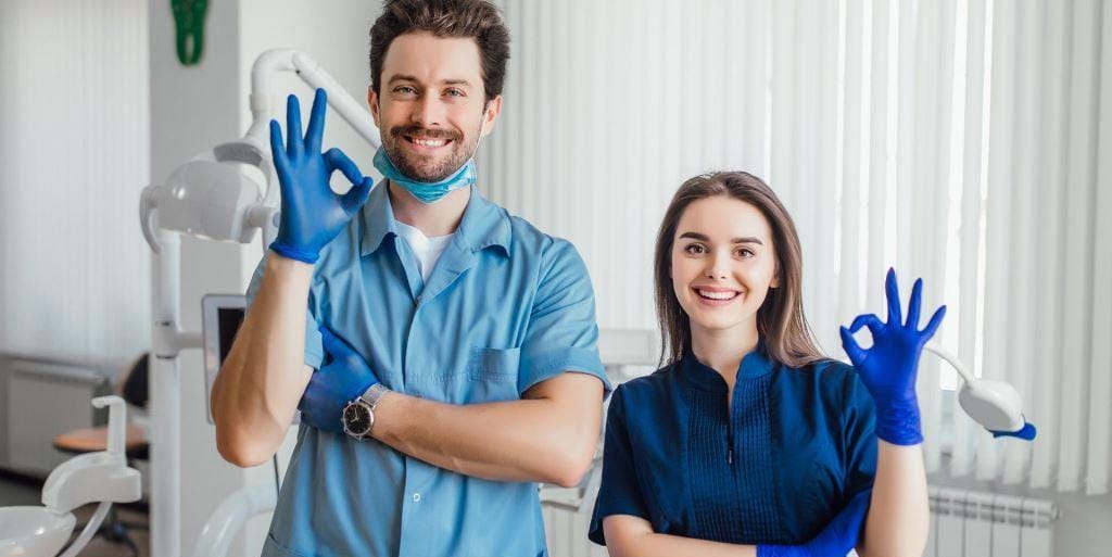 Two people in dental uniforms smiling and making an OK gesture