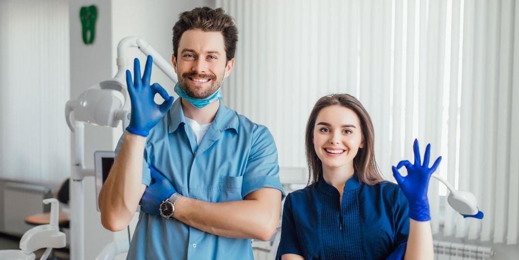 Two people in blue clinical clothing beside a dental chair
