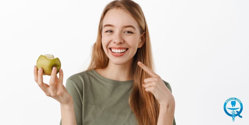 A woman holding an apple and pointing towards her teeth